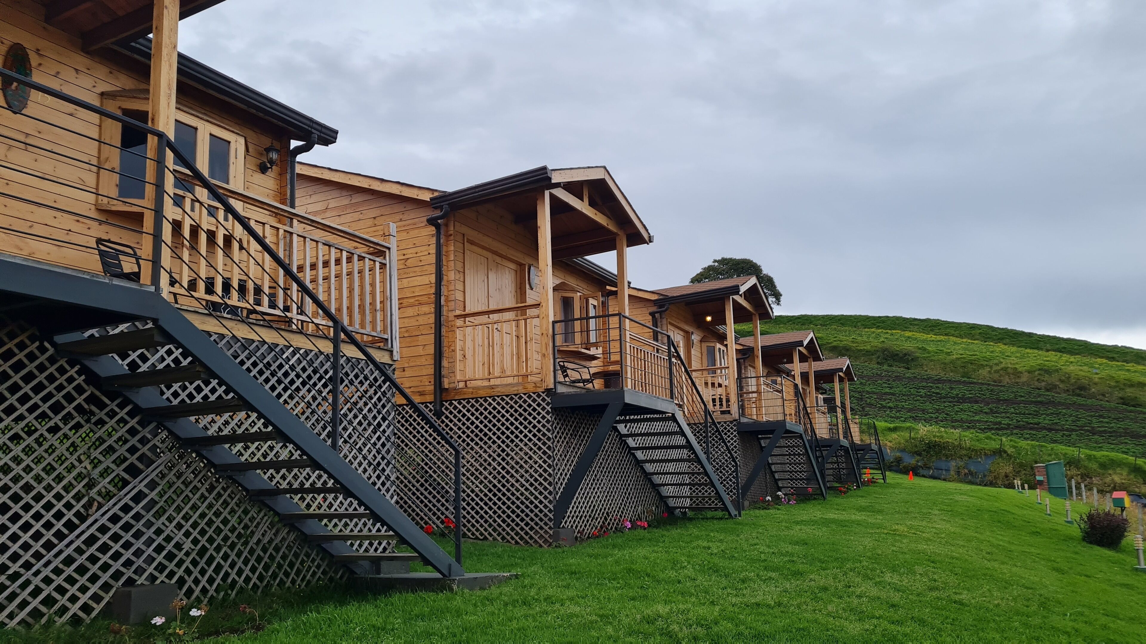 A row of wooden cabins with external staircases and a green hillside in the background.
