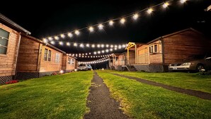 A row of wooden cabins with a paved path leading to a covered area, string lights overhead, and cars parked in front. - Cabañas & Mirador Secret Garden Costa R. (Potrero Cerrado)