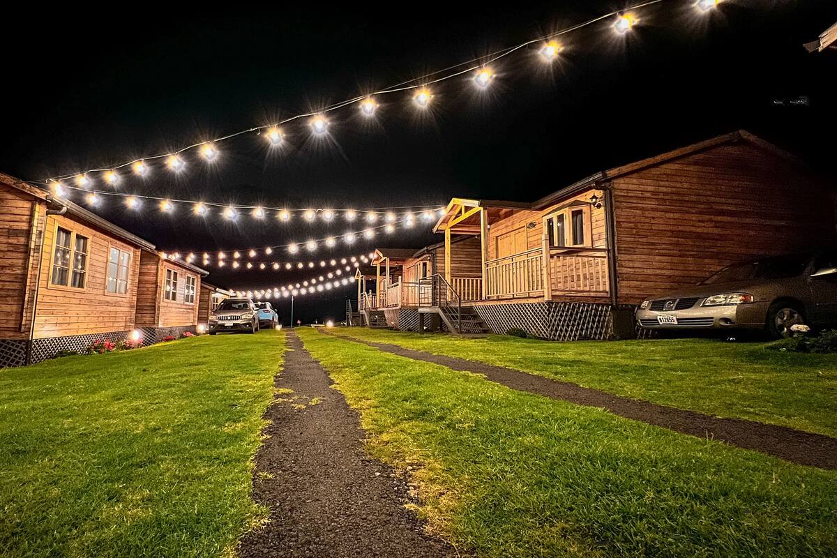 A row of wooden cabins with a paved path leading to a covered area, string lights overhead, and cars parked in front.
