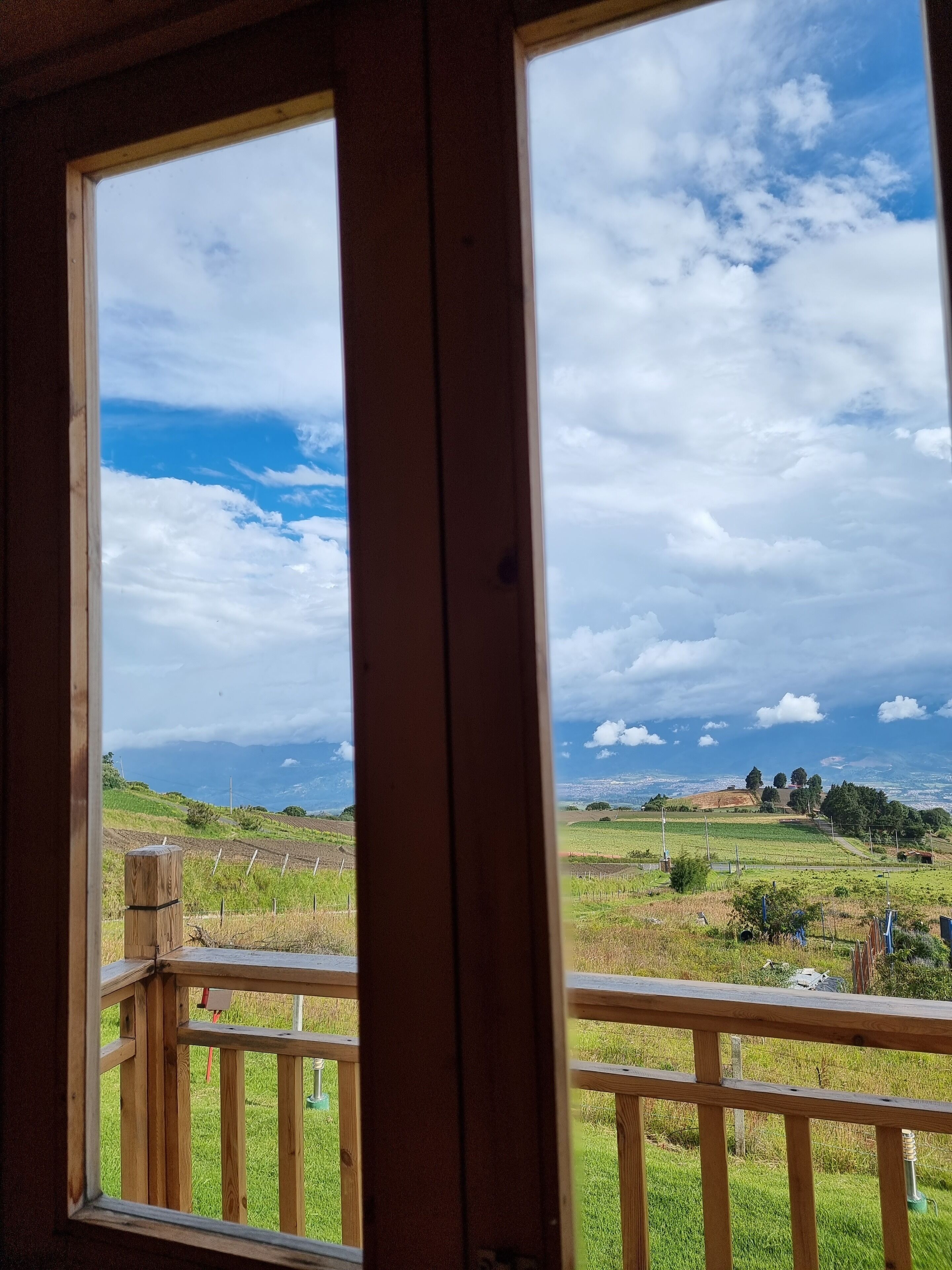 Vista desde un balcón de madera con barandilla, sobre un paisaje verde con campos y colinas distantes bajo un cielo parcialmente nublado.