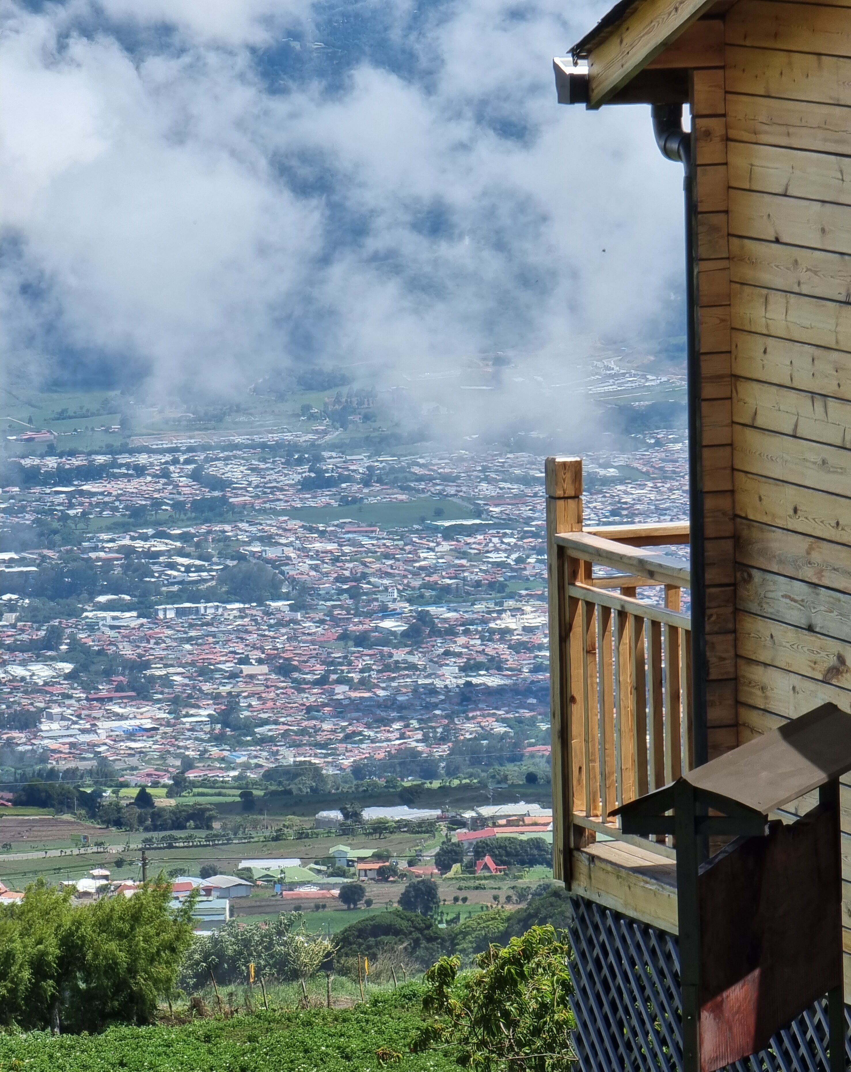 Ein hölzerner Balkon mit Blick auf eine Stadt und die umliegende Landschaft, Wolken am Himmel.