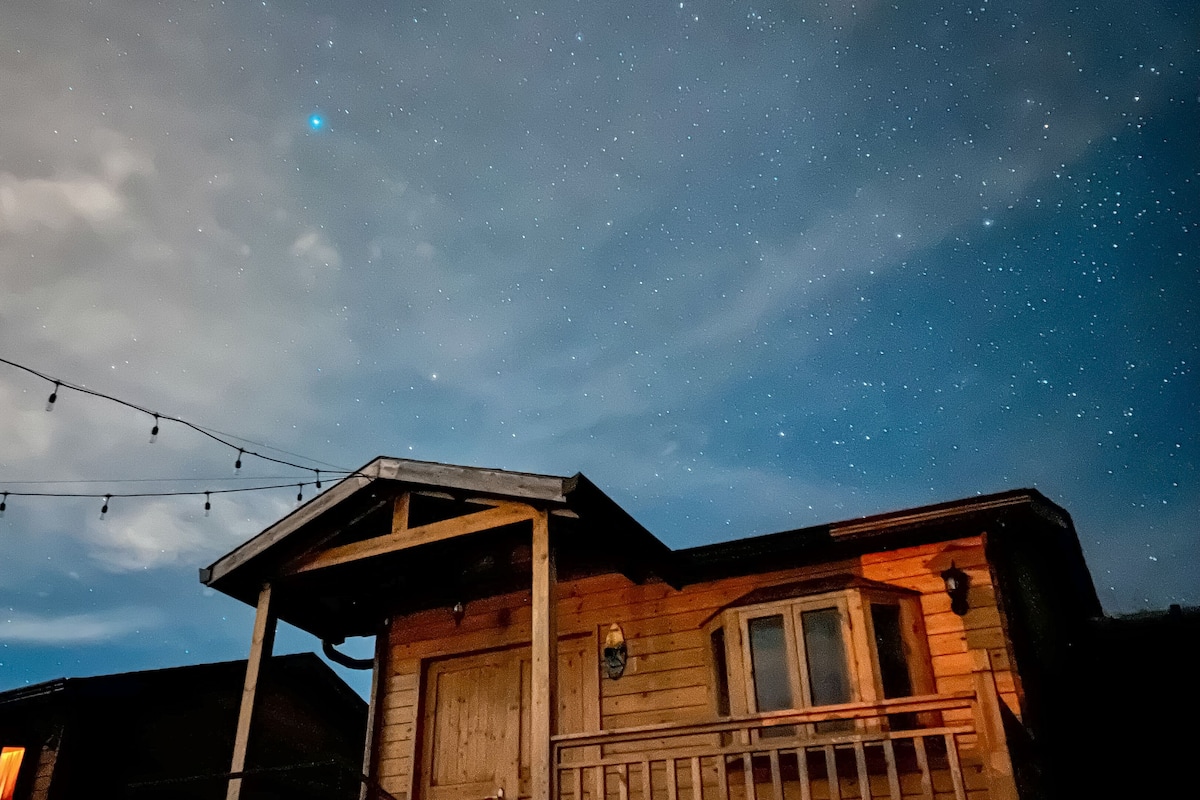 A wooden cabin with a porch and string lights against a starry night sky.