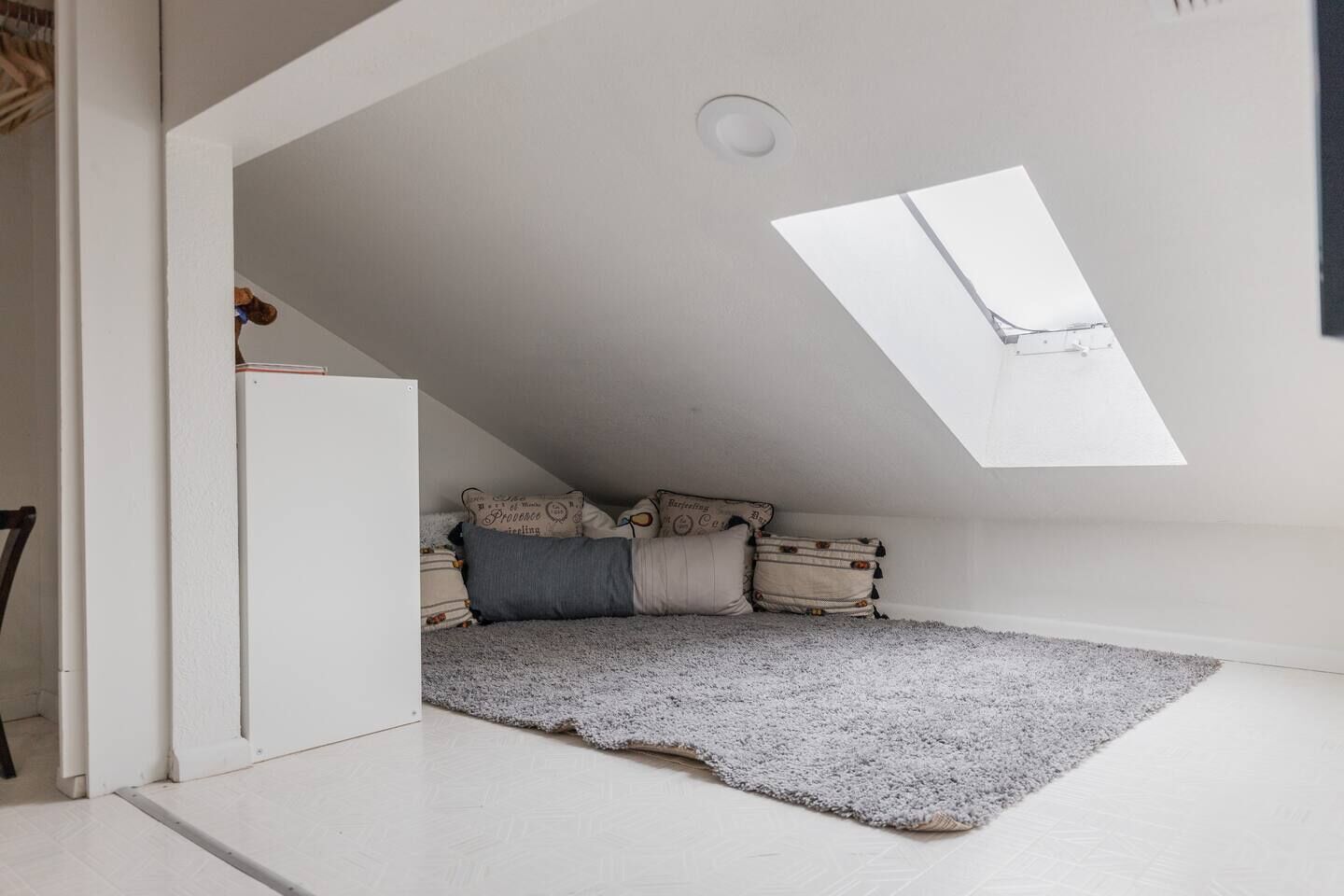 Une petite pièce avec un plafond en pente et une lucarne, un tapis gris et un lit recouvert de plusieurs coussins.