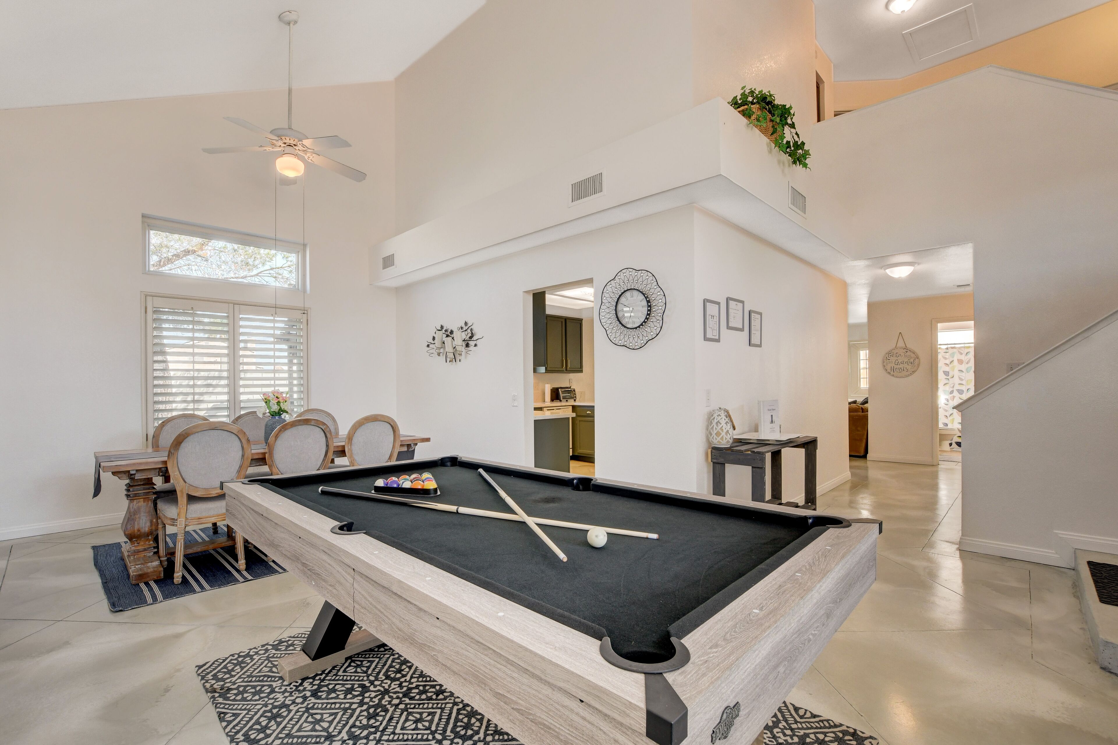 A pool table in a spacious room with a dining area, a ceiling fan, and a staircase leading to an upper level.