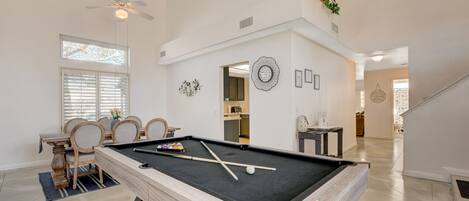 A pool table in a spacious room with a dining area, a ceiling fan, and a staircase leading to an upper level.