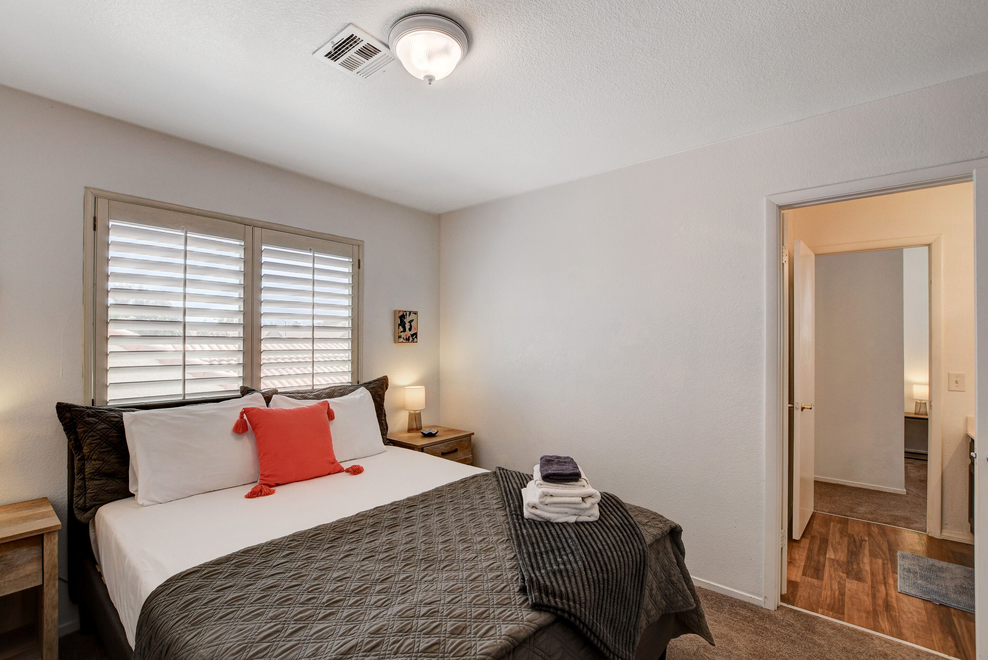 Une chambre à coucher comprenant un lit, une table de chevet, une lampe, une fenêtre avec des stores et une porte donnant sur une autre pièce.