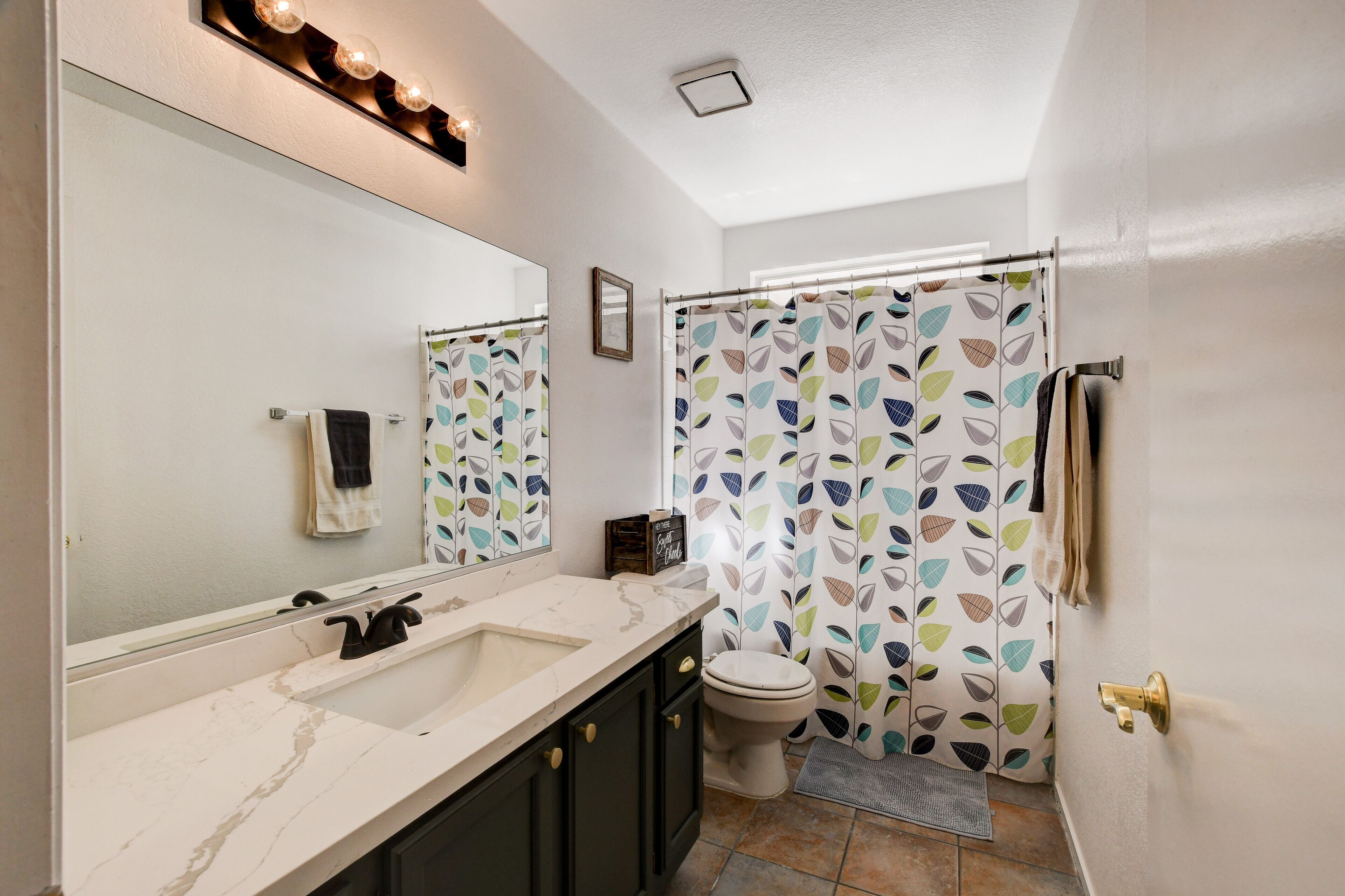 Un baño con lavamanos blanco, muebles oscuros, inodoro y cortina de ducha con motivos de hojas.