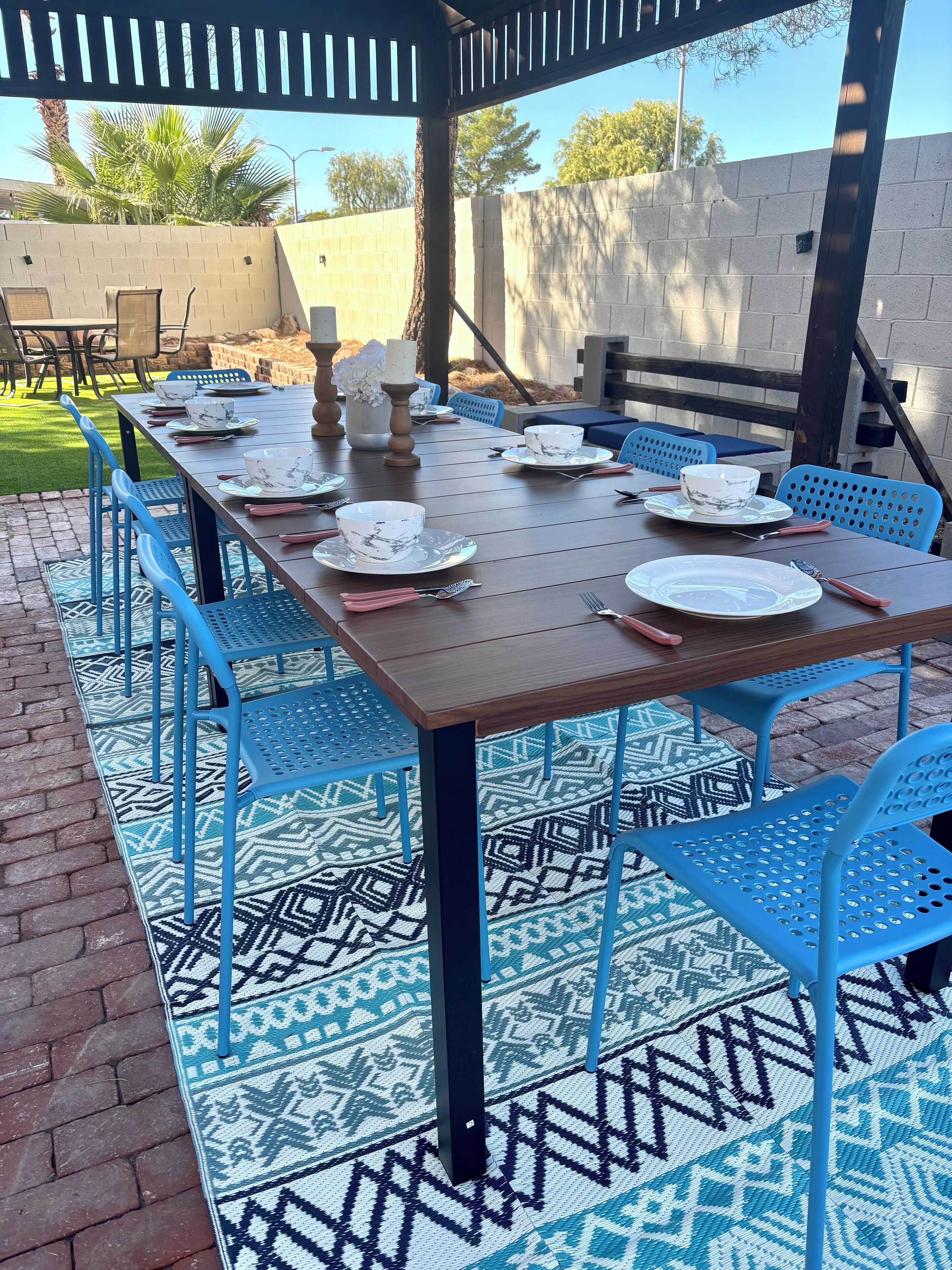 Eine Terrasse im Freien mit einem Holztisch, gedeckten Stellen zum Essen, blauen Stühlen und einem gemusterten Teppich.