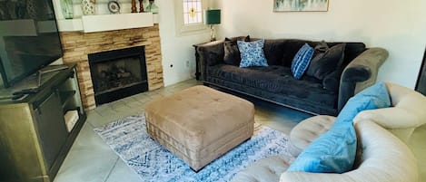 Una sala de estar con chimenea, un sofá, un reposapiés y un televisor.