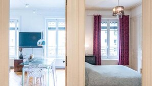 A hotel room with a wooden herringbone floor, a bed with a dark headboard, a chandelier, and a view of the city through large windows. - 128 Thiers (Villeurbanne)