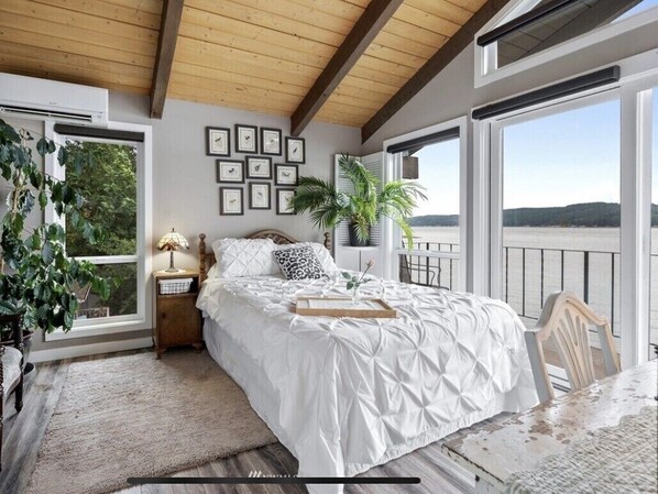 Un dormitorio con cama, mesita de noche, lámpara, ventana con vista al agua y una planta.