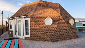 Una estructura de domo geodésico con revestimiento de madera, una puerta corrediza de vidrio y una terraza con tablones de madera de colores.