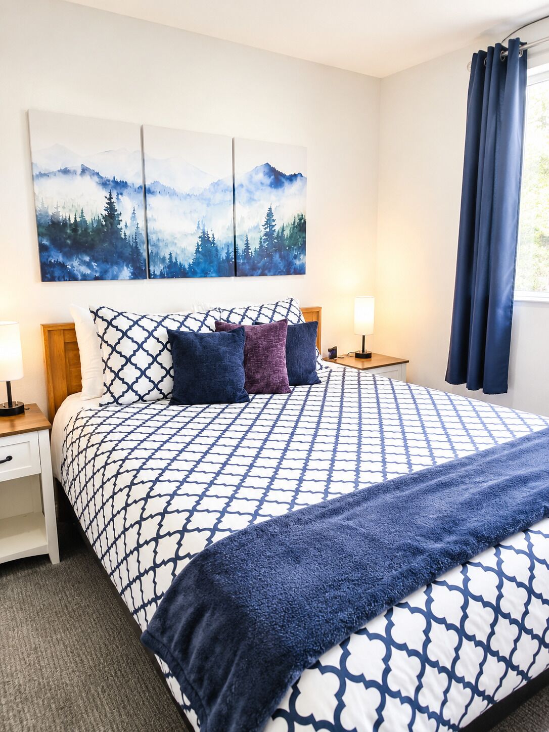 A bedroom with a large bed, a nightstand, a lamp, and a painting of a forest on the wall.