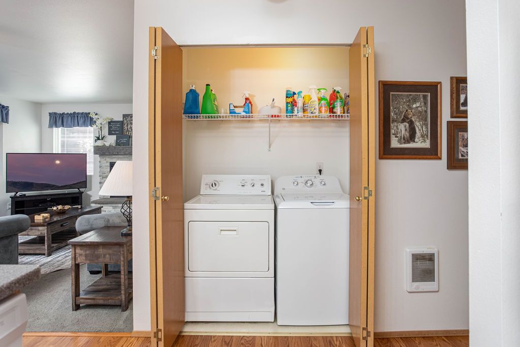 A laundry room with two white front-loading washers and dryers, a shelf with cleaning supplies, and a television in the adjacent living area.