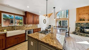 A spacious kitchen with granite countertops, wooden cabinets, a central island, pendant lighting, a stone fireplace, and large windows. - 11 Beds, Walk to Pool and Hot Tub, Game Room (Truckee)