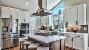 A modern kitchen with stainless steel appliances, a large island with stools, and a view of the outdoors through a large window. - Mountain Base Camp: Hot Tub, A/C, Dog Friendly (South Lake Tahoe)