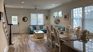 A spacious living room with a blue sofa, dining table, and chairs, a staircase, and a large window with blinds. - Mermaid Place is only 5 min walk to the beach. (Seaside Heights)