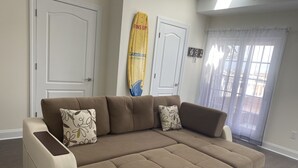 A modern living room with a sectional sofa, two decorative pillows, a surfboard, and a sliding glass door leading to a balcony. - Mermaid Place is only 5 min walk to the beach. (Seaside Heights)