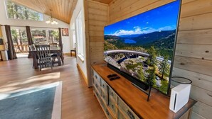 A wooden interior with a large TV displaying a scenic landscape, a dining area with chairs and a table, and a wooden cabinet with drawers.