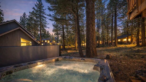 A hot tub surrounded by trees, wooden cabins, and a clear sky.