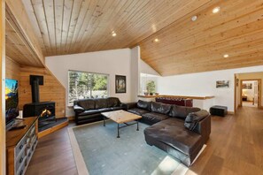 A modern living room with a fireplace, leather sofa, wooden ceiling, and large windows.