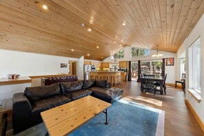 A spacious living area with a wooden ceiling, a black leather sofa, a wooden coffee table, and a kitchen in the background.