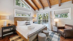 Ein Schlafzimmer mit einem großen Bett, Holzbalkendecke, zwei Fenstern, einem Nachttisch, einem Sessel und einem kleinen Tisch.
