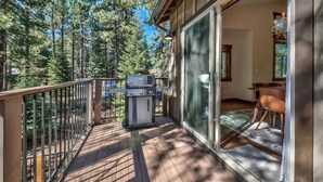A wooden deck with a grill, a glass door leading to a dining area, and a forested backdrop. - New Tahoe Cabin with Hot Tub Games and EV Charger (South Lake Tahoe)