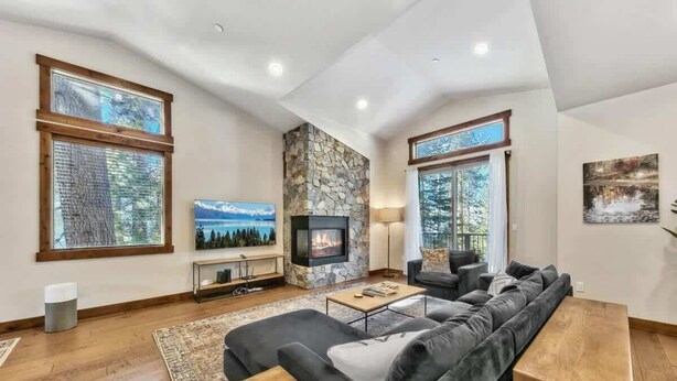 A modern living room with a stone fireplace, large windows, a sofa, and a coffee table. - New Tahoe Cabin with Hot Tub Games and EV Charger (South Lake Tahoe)
