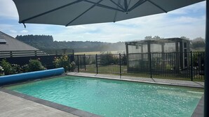 A swimming pool with a covered area, a fenced garden, and a distant view of trees and hills. - Hidden Valley Getaway (Martinborough)