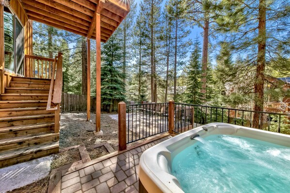 A wooden cabin with a hot tub, wooden stairs, and a fenced outdoor area surrounded by tall trees.