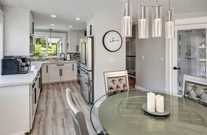Uma cozinha moderna com armários brancos, uma mesa de vidro redonda com velas, luminárias pendentes e um relógio na parede.