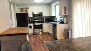 A compact kitchen with white cabinets, stainless steel appliances, and a wooden dining table. - Cozy 3 Bedroom Unit (Olympia)