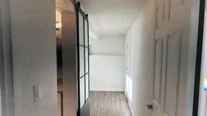 A narrow hallway with a sliding glass door leading to a room. The hallway has a wooden floor and white walls. - New! Charming 4-Bedroom House (Des Moines)