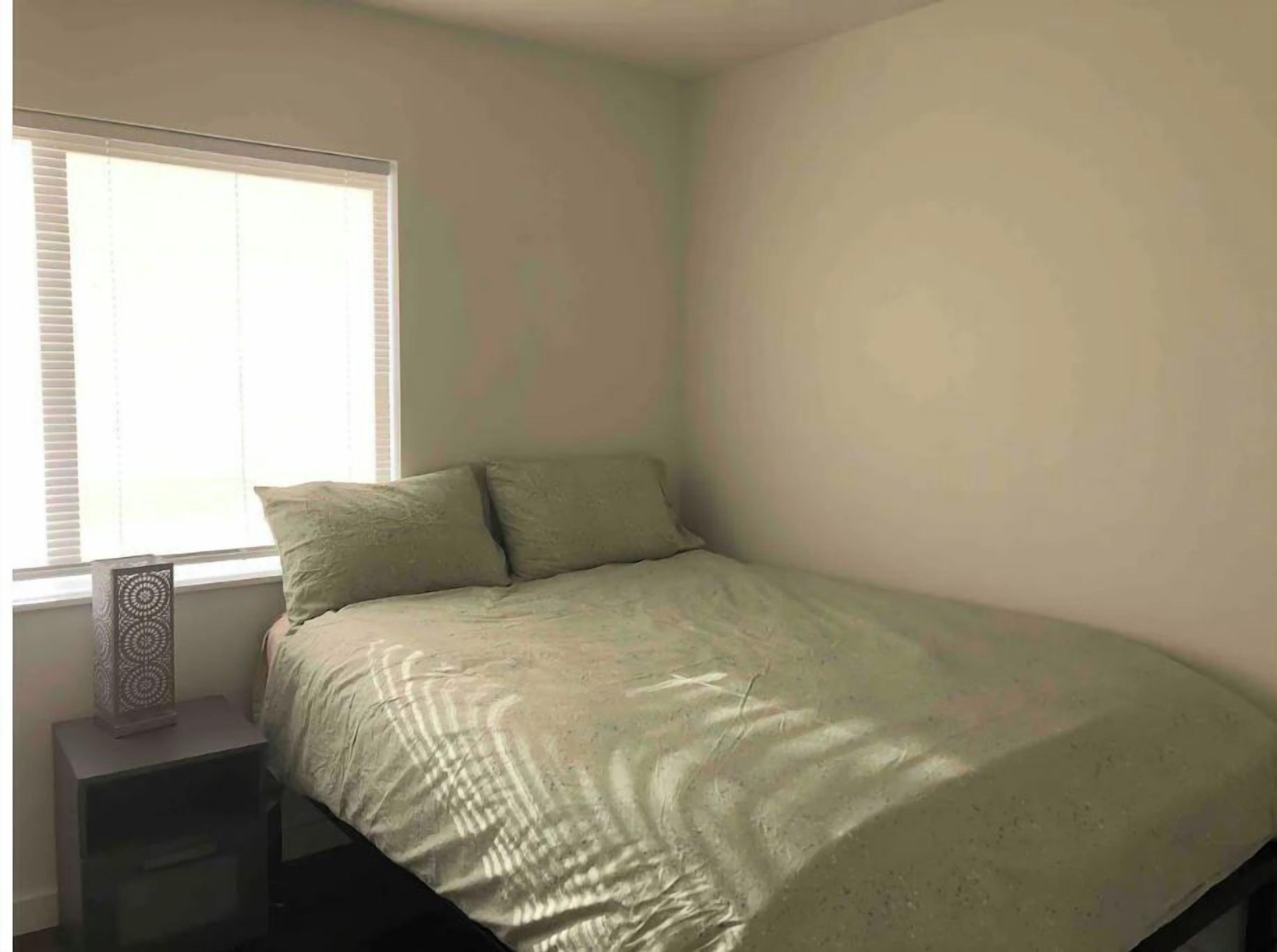 A small, single-bed room with a window, a bedside table, and a wall-mounted light fixture.