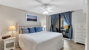 Un dormitorio con una cama grande, dos mesitas de noche con lámparas, un ventilador de techo, vista a través de un ventanal y un aparador blanco.