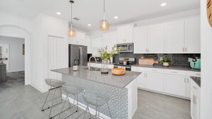 Une cuisine moderne avec des meubles blancs, un îlot central, des tabourets de bar, des luminaires suspendus et un four à micro-ondes.