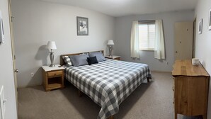 Une chambre à coucher avec un lit, deux tables de chevet avec des lampes, une commode en bois, une fenêtre avec des rideaux et un tableau encadré au mur.