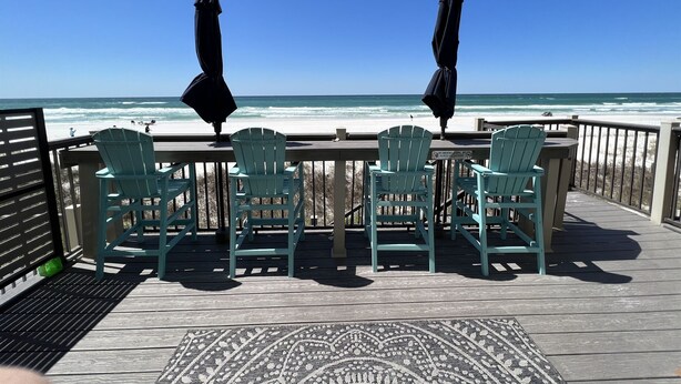 A wooden deck with four green Adirondack chairs and two black umbrellas, overlooking a beach with waves.