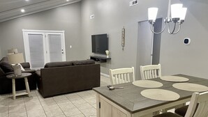 Ein modernes Wohnzimmer mit einer Couch, einem Esstisch mit Platzsets, einem Fernseher und einer Deckenleuchte.