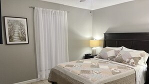 Ein Schlafzimmer mit einem Bett, einem Nachttisch, einer Lampe, einem gerahmten Bild an der Wand und einem Fenster mit weißem Vorhang.