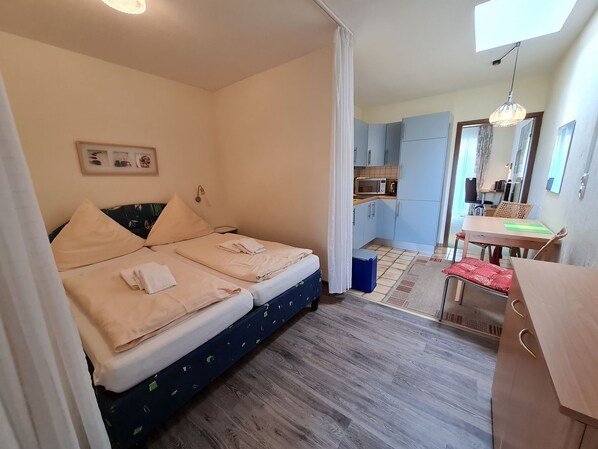 A bedroom with a bed, a bedside table, a small desk with a chair, a kitchenette, and a bathroom.