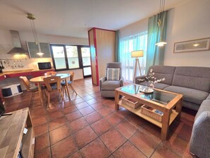 Ein Wohnzimmer mit einer Couch, einem Couchtisch, einem Esstisch mit Stühlen, einer Küchenzeile mit einer Mikrowelle und einem Fenster mit Vorhängen.