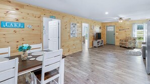 A cabin interior with a wooden interior, a dining area with white chairs, a table with a vase, and a sign that reads "What happens at the lake stays at the lake." - Relaxing, family friendly lodge by the lake. (Ten Mile)