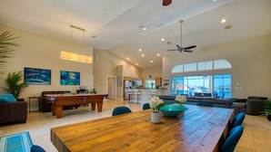 A spacious living room with a wooden dining table, blue chairs, a pool table, and a large window offering a view of the outdoors. - Intervillas Florida - Villa Manatee (Cape Coral)