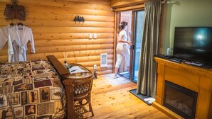 Une chambre de style rustique, aménagée dans une cabane en rondins, avec un lit, une télévision, une cheminée et une porte-fenêtre coulissante donnant sur l’extérieur.