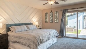 Un dormitorio con cama, mesita de noche, ventilador de techo y vistas al exterior a través de un ventanal corredizo.