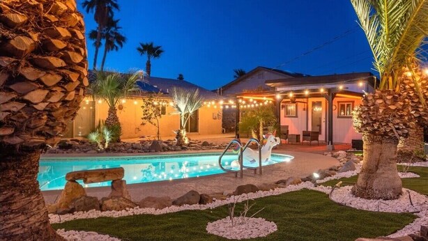 Un área de piscina con un banco de piedra, palmeras y luces colgantes.
