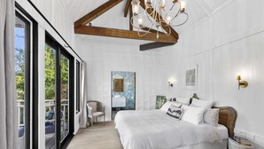 A bedroom with a large bed, a wooden ceiling beam, a chandelier, and a view through a sliding glass door.