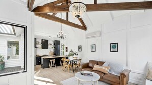 A spacious living room with a brown leather sofa, a round coffee table, and a dining area with wooden chairs. A kitchen with black cabinets and a chandelier is visible in the background.